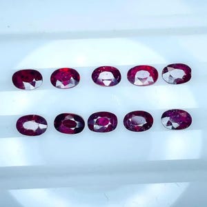 Puede incluir: Diez piedras preciosas de color rojo intenso con corte ovalado, dispuestas en dos filas horizontales de cinco. Cada piedra presenta una superficie facetada que refleja la luz, mostrando un tono carmesí rico y saturado con claridad interna.