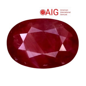 Puede incluir: Una piedra preciosa de color rojo intenso con corte ovalado y superficie facetada que refleja la luz. Sobre la piedra, el texto AIG American International GemLab aparece en letras rojas y negras sobre un fondo blanco liso.