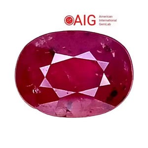 Puede incluir: Una gema de rubí de corte ovalado con un patrón de estrella en el centro. La piedra de color rojo intenso se encuentra sobre un fondo blanco. El logotipo "AIG American International GemLab" está en la esquina superior izquierda.