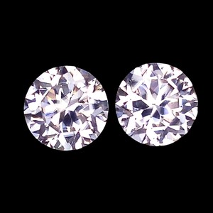 May include: Two round-cut, clear, faceted gemstones displayed against a solid black background. Each stone features a brilliant, symmetrical pattern of light-reflecting facets that create a sparkling, multi-dimensional appearance with white and light gray tones.