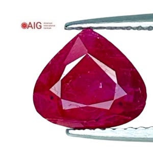 May include: A faceted, pear-shaped ruby gemstone, held by tweezers against a white background. The ruby is a deep, vibrant red color, with multiple facets reflecting light. The AIG logo is visible in the upper left corner.