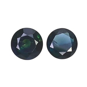 Peut inclure: Deux pierres précieuses rondes à facettes avec une teinte vert foncé et bleu profond. Chaque pierre présente une taille circulaire précise avec de multiples facettes triangulaires qui reflètent la lumière, créant un aspect poli et profond.