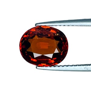 Puede incluir: Una gema ovalada y facetada de color marrón rojizo intenso. La gema está sujeta por unas pinzas plateadas sobre un fondo blanco. Las facetas crean un patrón en forma de estrella en el centro de la piedra.