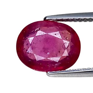 Könnte beinhalten: Ein oval geschliffener Rubin-Edelstein mit tiefroter Farbe und facettierter Oberfläche, gehalten von einer silberfarbenen Metallpinzette. Der Stein weist interne Einschlüsse und ein poliertes Finish auf und zeigt eine klassische Form.