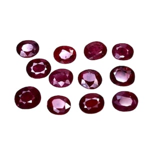 May include: An assortment of oval-shaped, deep red gemstones, possibly rubies, are scattered against a white background. The gems vary slightly in color and clarity, showcasing natural variations. The polished stones are ready for jewelry making.
