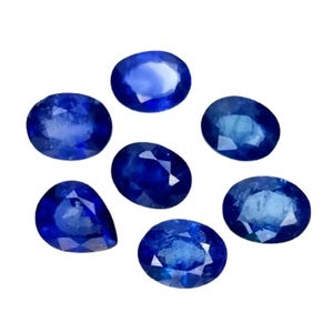 May include: A collection of seven deep blue sapphires in various shapes, including oval and pear cuts, against a white background. The gemstones display a rich, saturated color and a polished surface, reflecting light.