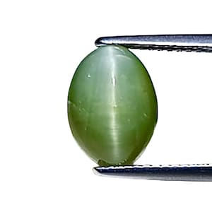 May include: A light green, oval-shaped gemstone, held by tweezers against a white background. The gemstone has a smooth, polished surface and a cat's eye effect, with a band of light running across its center. The gem is likely a Chrysoberyl.