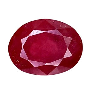 May include: A faceted, oval-shaped ruby gemstone. The gem is a deep, saturated red color with a brilliant, polished surface. The facets create a starburst pattern in the center of the stone, reflecting light. The ruby is set against a white background.