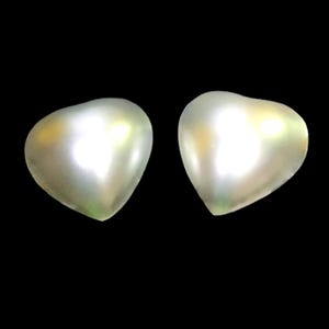 Puede incluir: Dos objetos nacarados en forma de corazón con un brillo luminoso. Los objetos están sobre un fondo negro, resaltando sus superficies lisas y reflectantes. Los corazones tienen una forma suave y redondeada, sugiriendo un tema romántico.