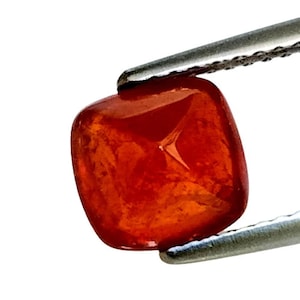 Puede incluir: Una gema pulida, de corte cuadrado, de color rojo anaranjado, sujeta con pinzas plateadas. La gema tiene esquinas redondeadas y una superficie brillante, que refleja la luz. El fondo es blanco, resaltando el color y la claridad.