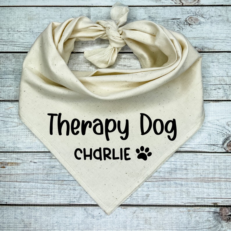 Może przedstawiać: Biała bawełniana bandana dla psa z czarnym tekstem, kt&oacute;ry brzmi "Therapy Dog CHARLIE" z odciskiem łapy.