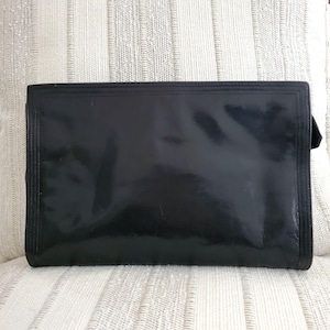 May include: A rectangular black clutch with a glossy finish. The clutch has a simple design with stitched edges and a small loop on the side. The bag is resting on a striped, off-white surface.