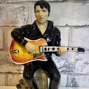 May include: A ceramic figurine of Elvis Presley playing a guitar, sitting on a ledge with a small dog beside him. The figurine is painted in realistic colours, with Elvis wearing a black leather jacket and black trousers. The guitar is a brown and yellow electric guitar with a black pickguard.