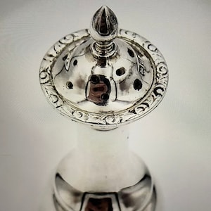 Könnte beinhalten: Silberner Pfefferstreuer mit einem gewölbten Deckel und einem kleinen Knopf oben. Der Deckel hat einen dekorativen Rand und Löcher, durch die Pfeffer gelangen kann.