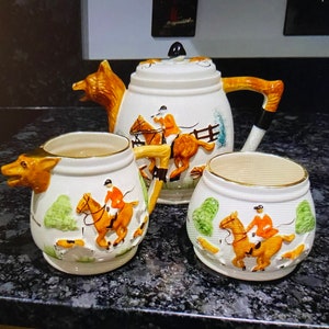 May include: A white ceramic teapot and two mugs with fox head handles. The teapot and mugs are decorated with a hunting scene featuring riders on horses and hounds.