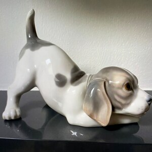 May include: A porcelain figurine of a white and gray puppy with brown eyes, lying on its belly with its tail curled up. The puppy is looking to the right.