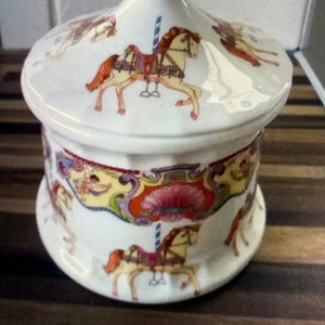 Op de afbeelding: Witte keramische pot met een deksel met een carrouselontwerp. Het deksel heeft een kleurrijk carrouselscène met paarden, een paal en een luifel. De pot heeft een vergelijkbaar ontwerp met een kleurrijk carrouselscène en een roze bloem.