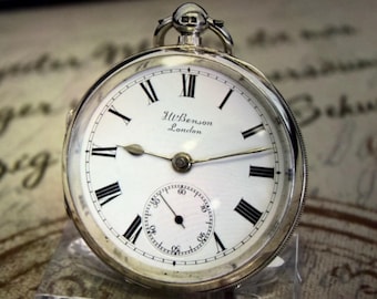 May include: A silver pocket watch with a white face and black Roman numerals. The watch face includes a smaller seconds dial and the text "J.W. Benson London". The watch has a silver crown and a loop for a chain.