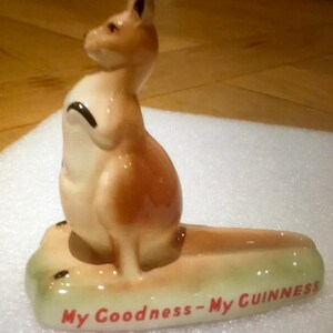 K&ouml;nnte beinhalten: Eine Keramik-K&auml;nguru-Figur in Beige- und Cremet&ouml;nen mit einem braunen Fleck auf dem R&uuml;cken. Das K&auml;nguru steht auf einem hellgr&uuml;nen Sockel mit dem Text "My Goodness - My GUINNESS" in Rot.