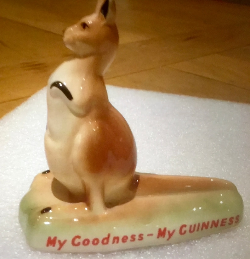 K&ouml;nnte beinhalten: Eine Keramik-K&auml;nguru-Figur in Beige- und Cremet&ouml;nen mit einem braunen Fleck auf dem R&uuml;cken. Das K&auml;nguru steht auf einem hellgr&uuml;nen Sockel mit dem Text "My Goodness - My GUINNESS" in Rot.