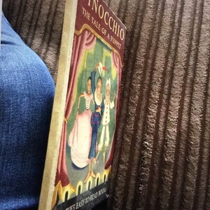 May include: A vintage copy of "Pinocchio: The Tale of a Puppet" by Blackie's Easy-to-Read Books. The cover features a colorful illustration of the puppet show with characters in costume. The book is held against a blue denim background.