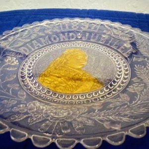 Può includere: Un piatto in vetro trasparente con bordo ondulato presenta un'immagine dorata della regina Vittoria al centro. Il piatto è decorato con un motivo floreale e il testo "1897 DIAMOND JUBILEE".