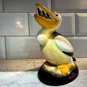 May include: A ceramic pelican figurine with a yellow beak and feet, holding several small bottles in its mouth. The pelican has black and white wings and a black base with the word "Guinness" in red.