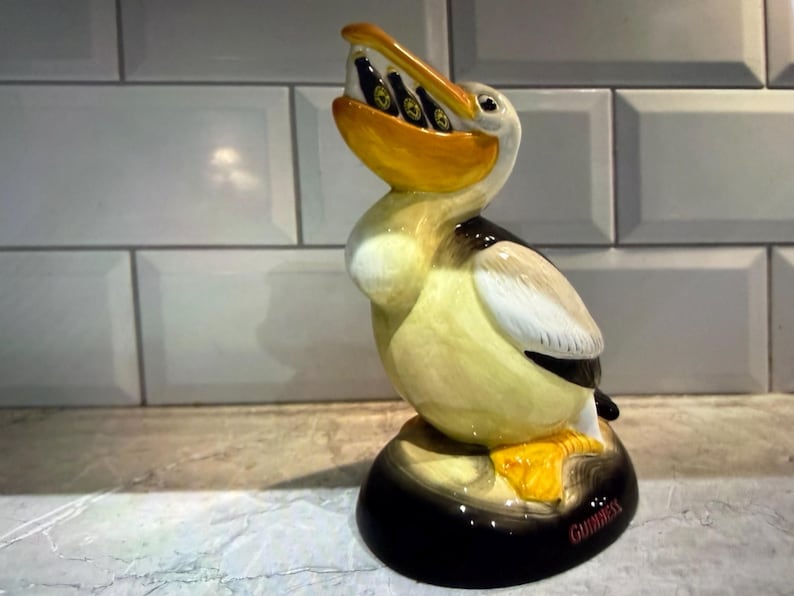 May include: A ceramic pelican figurine with a yellow beak and feet, holding several small bottles in its mouth. The pelican has black and white wings and a black base with the word "Guinness" in red.