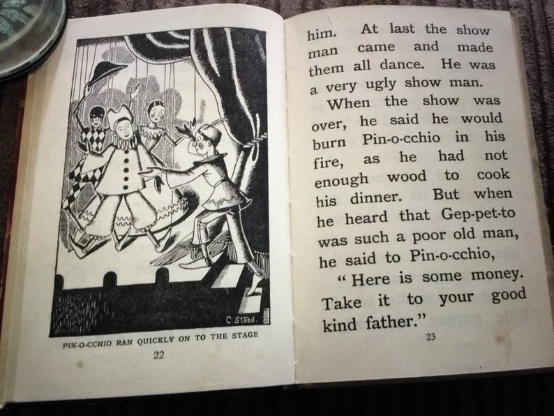 May include: An open book with a black and white illustration of a scene from Pinocchio. The illustration shows characters on a stage, with text that reads "Pin-occhio ran quickly on to the stage".