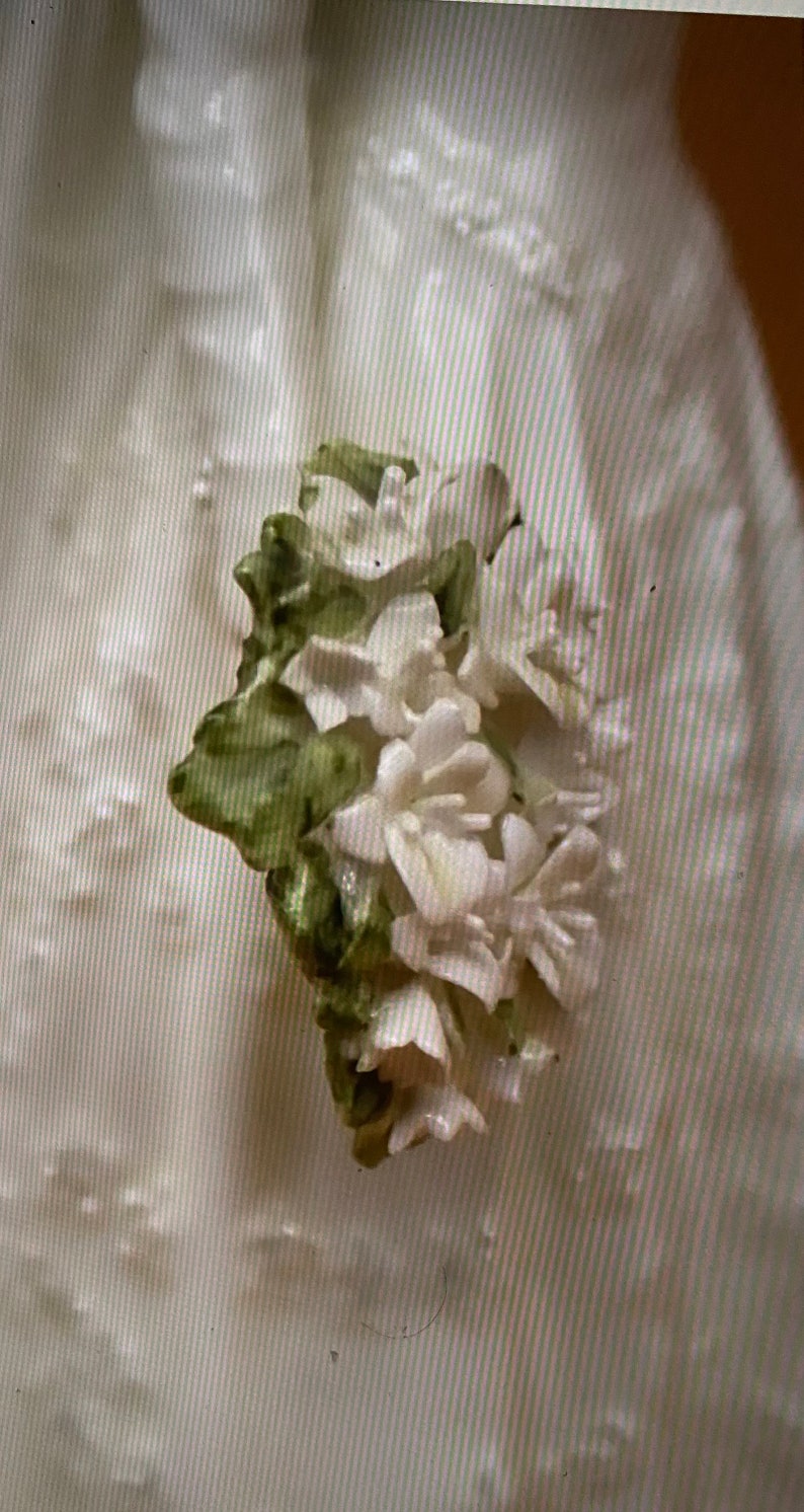 May include: A close-up of a white porcelain flower with green leaves. The flower is delicate and intricate, with a realistic appearance.
