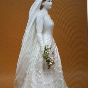 May include: A white porcelain figurine of a bride wearing a long white gown with a veil and holding a bouquet of white flowers.