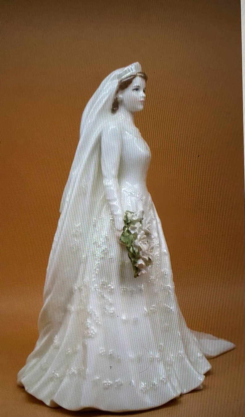 May include: A white porcelain figurine of a bride wearing a long white gown with a veil and holding a bouquet of white flowers.