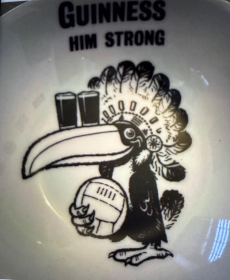 May include: White ceramic bowl with a black and white illustration of a toucan wearing a Native American headdress and holding a volleyball. The bowl has the text "GUINNESS HIM STRONG" in black.