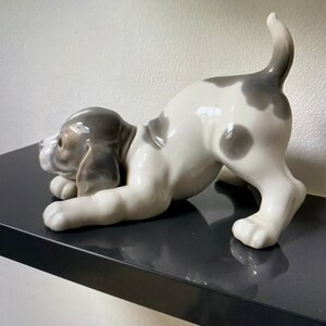May include: A white and gray porcelain figurine of a dog in a playful pose. The dog is lying on its belly with its head turned to the side and its tail curled up. The figurine is made of high-quality porcelain and is a beautiful addition to any home.