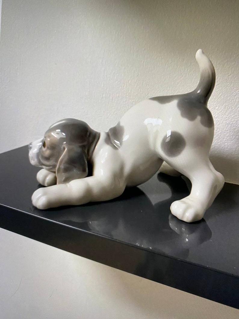 May include: A white and gray porcelain figurine of a dog in a playful pose. The dog is lying on its belly with its head turned to the side and its tail curled up. The figurine is made of high-quality porcelain and is a beautiful addition to any home.