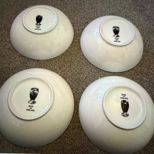 May include: Four white ceramic bowls. Each bowl has a circular base with a black and white graphic of a glass and the words "MADE IN ENGLAND". The bowls are arranged on a textured surface.