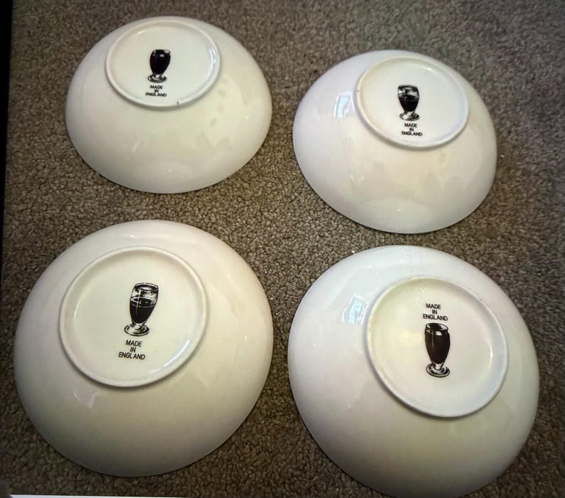 May include: Four white ceramic bowls. Each bowl has a circular base with a black and white graphic of a glass and the words "MADE IN ENGLAND". The bowls are arranged on a textured surface.