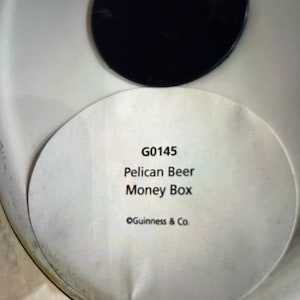 May include: A white money box with a black lid. The label reads "G0145 Pelican Beer Money Box" with "&copy;Guinness & Co." below. The money box is likely for collecting coins.