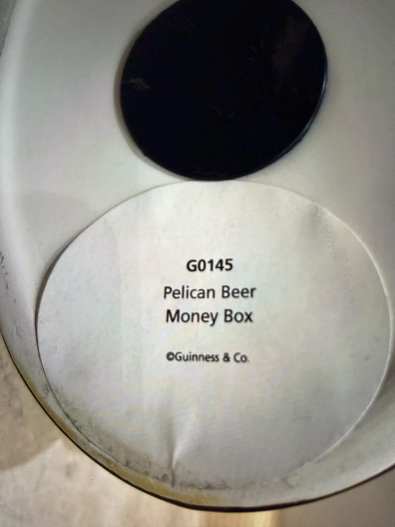 May include: A white money box with a black lid. The label reads "G0145 Pelican Beer Money Box" with "&copy;Guinness & Co." below. The money box is likely for collecting coins.