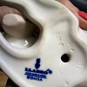 May include: White porcelain figurine with a blue and white Lladro logo and the text "DAISA" on the bottom. The figurine has a hole in the bottom and the number "1554" is visible.