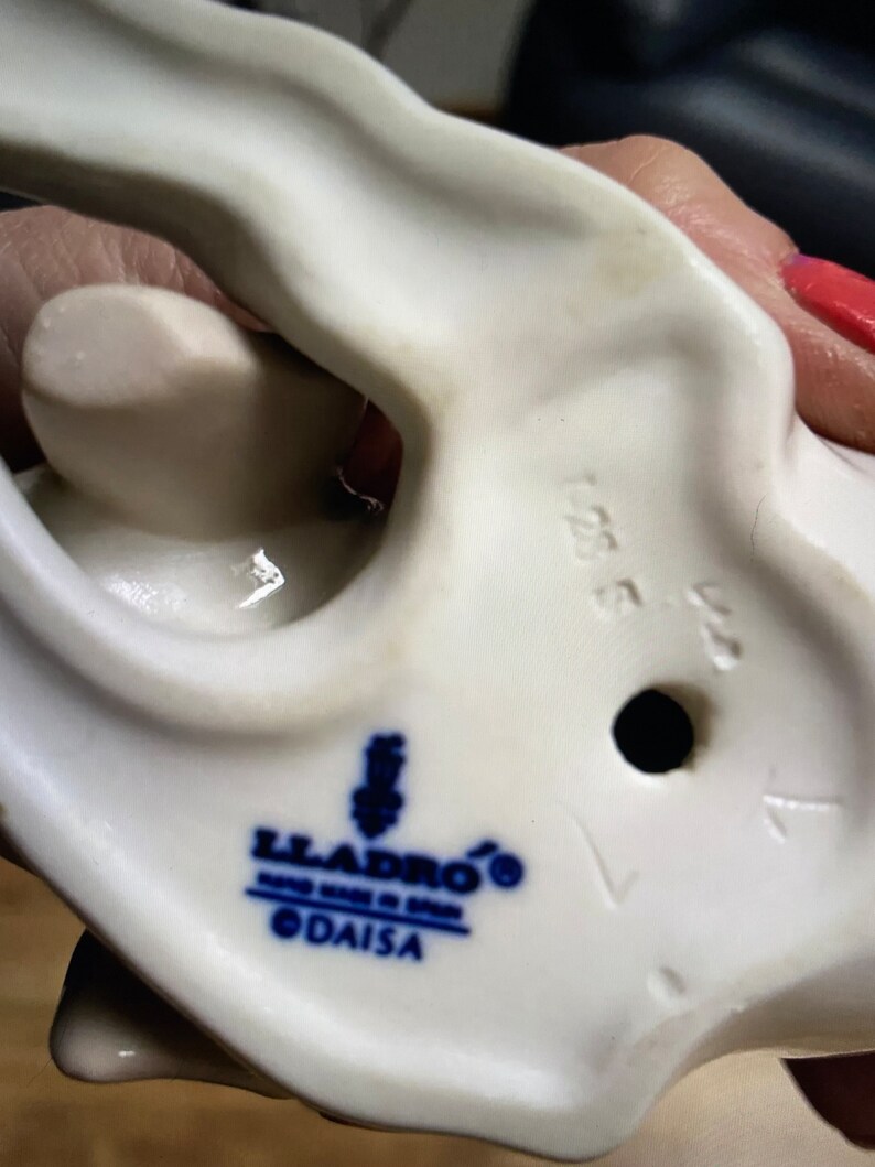 May include: White porcelain figurine with a blue and white Lladro logo and the text "DAISA" on the bottom. The figurine has a hole in the bottom and the number "1554" is visible.