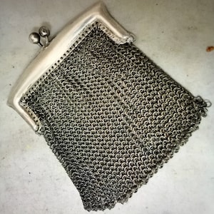 Puede incluir: Una cartera de monedas de malla plateada con un cierre de bisagra. La cartera está hecha de pequeños anillos de metal unidos.