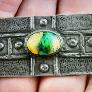 Puede incluir: Un broche de peltre con un diseño de esmalte verde y amarillo. El broche tiene una superficie texturizada y un diseño geométrico.