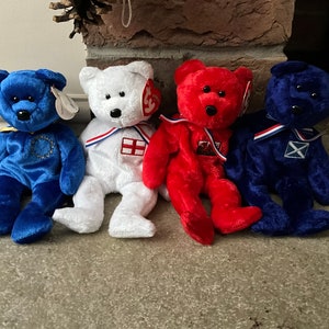 May include: Four plush teddy bears wearing different coloured outfits and hats. The bears are sitting on a grey surface. The bears are wearing outfits with the flags of England, Scotland, Wales, and Ireland.