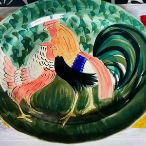 Könnte beinhalten: Eine ovale Keramikplatte mit grünem Hintergrund und drei darauf gemalten Hähnen. Die Hähne sind in den Farben Rosa, Orange, Schwarz und Blau bemalt. Die Platte eignet sich perfekt zum Servieren von Speisen oder als Dekorationsobjekt.