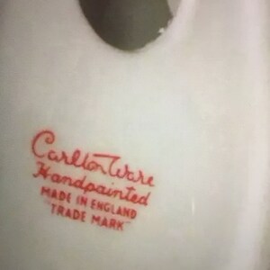 K&ouml;nnte beinhalten: Nahaufnahme eines wei&szlig;en Keramikobjekts mit einer dunklen &Ouml;ffnung. Der Text "Carlton Ware Handpainted Made in England Trade Mark" ist in roter Schrift gedruckt, was auf die Herkunft und Handwerkskunst des Artikels hinweist.