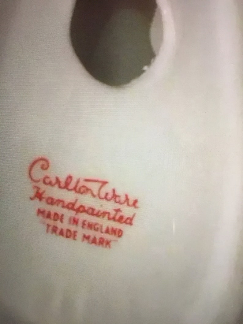 K&ouml;nnte beinhalten: Nahaufnahme eines wei&szlig;en Keramikobjekts mit einer dunklen &Ouml;ffnung. Der Text "Carlton Ware Handpainted Made in England Trade Mark" ist in roter Schrift gedruckt, was auf die Herkunft und Handwerkskunst des Artikels hinweist.