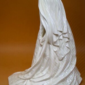 May include: White porcelain figurine of a bride wearing a flowing veil and dress. The figurine is posed with the bride's back to the viewer, and the veil is draped over her head and shoulders.