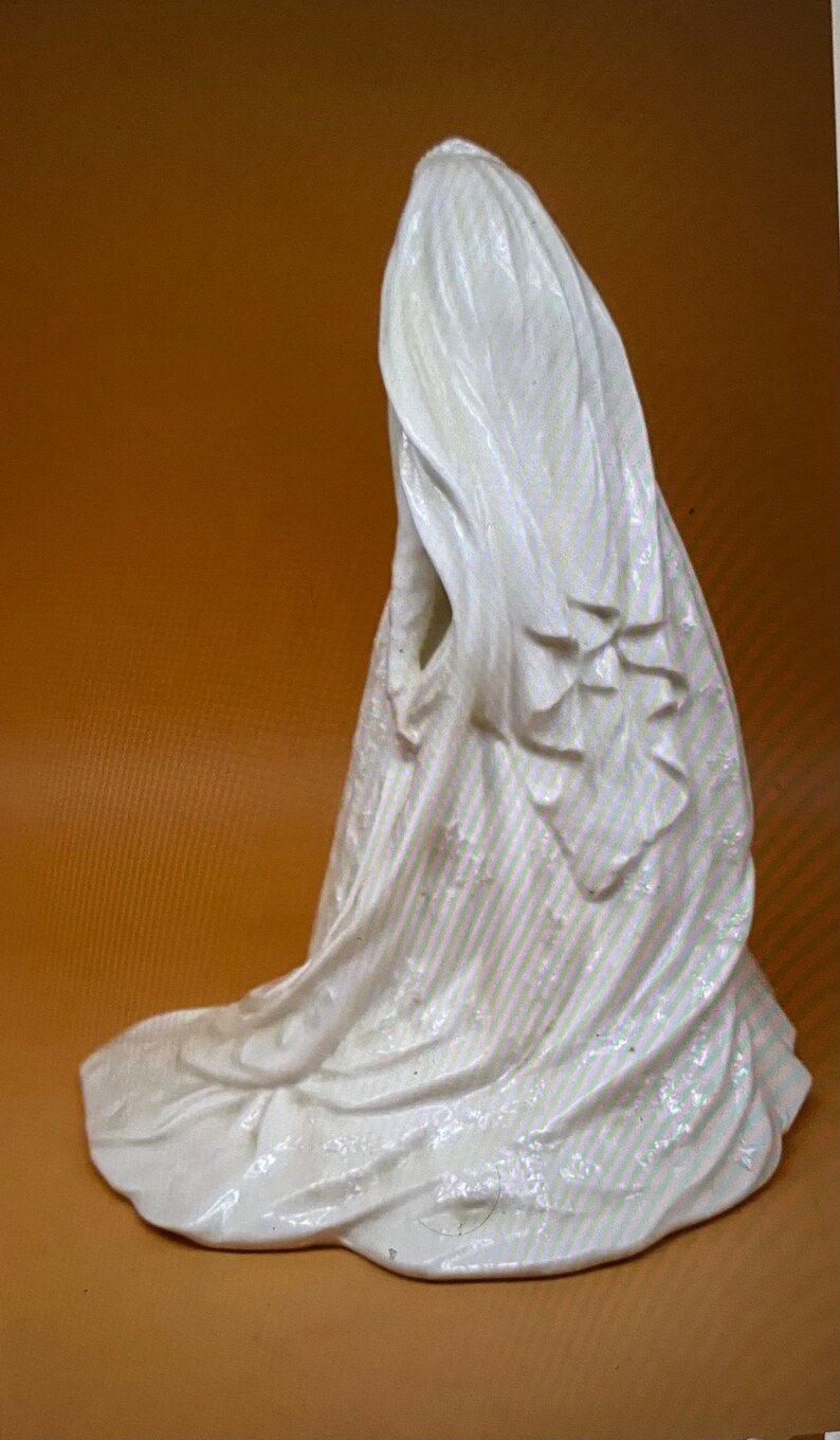May include: White porcelain figurine of a bride wearing a flowing veil and dress. The figurine is posed with the bride's back to the viewer, and the veil is draped over her head and shoulders.