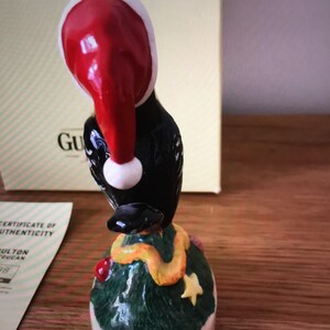 May include: A black bird figurine wearing a red Santa hat and a yellow scarf, standing on a green Christmas tree base with a pink bottom.